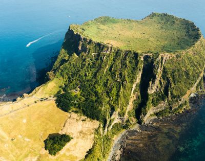 Aerial view of a volcano with steep, green cliffs and a flat grassy top, surrounded by bright blue ocean, exuding a tranquil and majestic vibe.