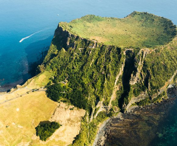 Aerial view of a volcano with steep, green cliffs and a flat grassy top, surrounded by bright blue ocean, exuding a tranquil and majestic vibe.