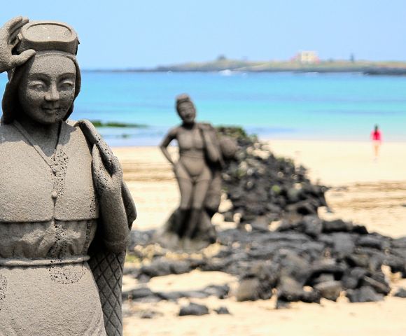 Stone statues of women on a sandy beach, with one in front smiling under a blue sky. Calm sea in the background; serene, tranquil atmosphere.