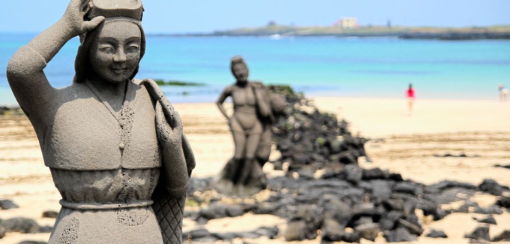 Stone statues of women on a sandy beach, with one in front smiling under a blue sky. Calm sea in the background; serene, tranquil atmosphere.