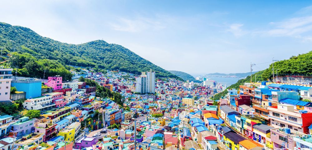 A vibrant hillside view of Gamcheon Culture Village in Busan, South Korea, showcasing colorful houses against a backdrop of mountains and ocean.