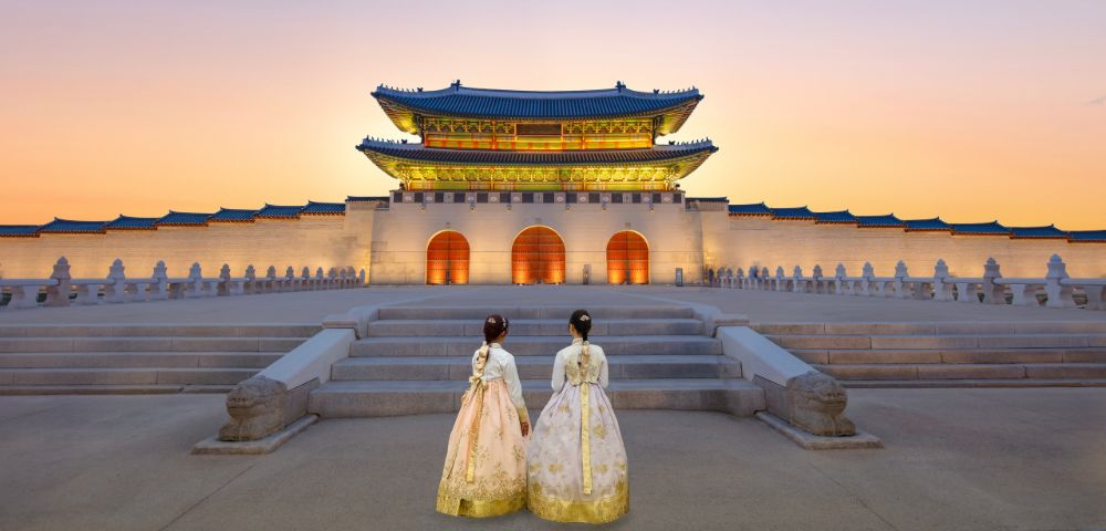 Two women in traditional hanbok admire the Gyeongbokgung Palace at sunset, with vibrant colors illuminating the historic architecture.
