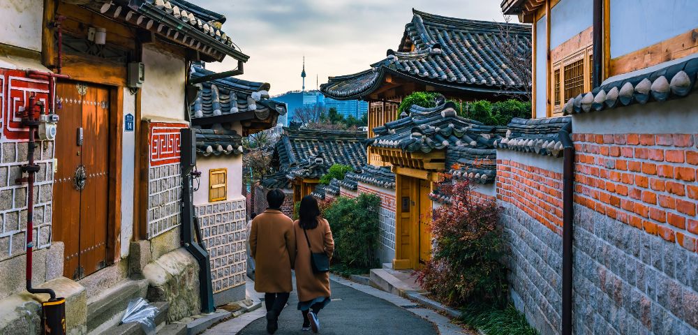 Two people walking side by side down a narrow street in a bustling Asian city, surrounded by shops and buildings.