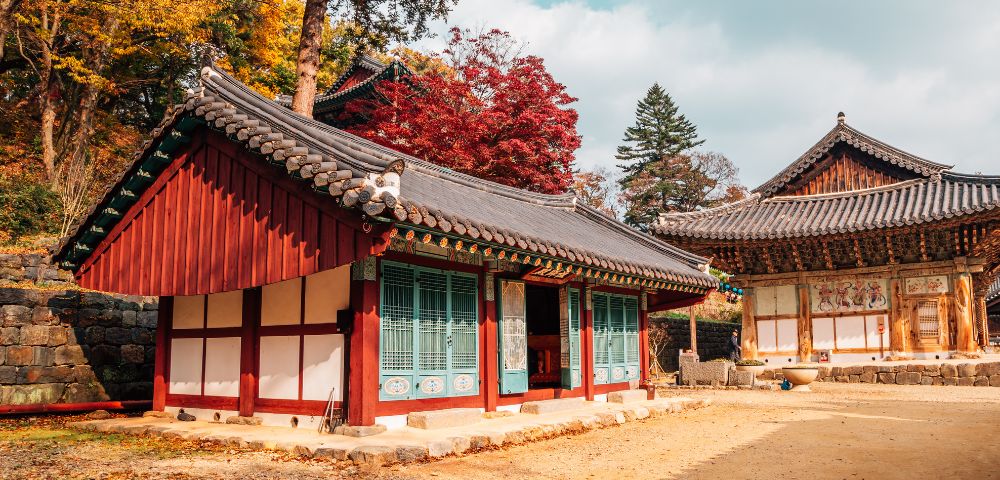 Traditional Korean buildings with colorful eaves and wooden shutters, surrounded by vibrant autumn foliage under a cloudy sky.