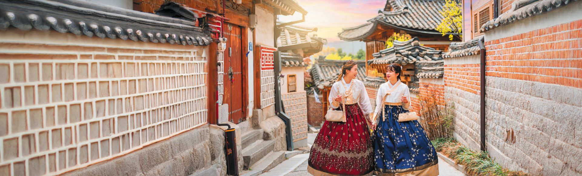Two women in traditional hanbok stroll through a charming, historical street lined with traditional Korean houses at sunset.