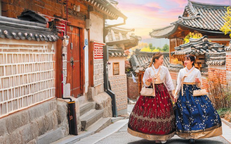 Two women in traditional hanbok stroll through a charming, historical street lined with traditional Korean houses at sunset.