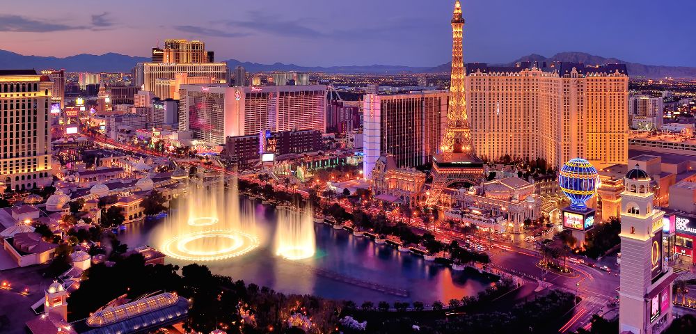 Aerial view of the Las Vegas Strip at dusk. Neon lights illuminate hotels and casinos, including a lit replica Eiffel Tower. A fountain show adds elegance.