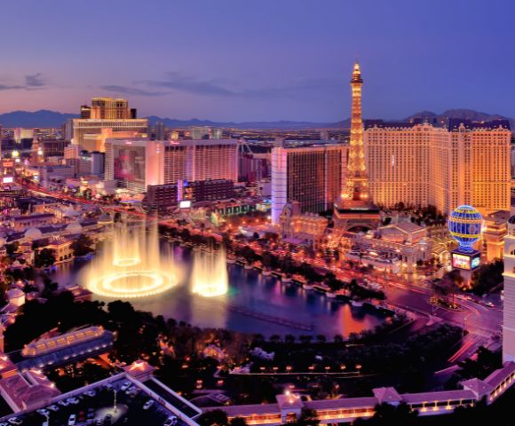 Aerial view of the Las Vegas Strip at dusk. Neon lights illuminate hotels and casinos, including a lit replica Eiffel Tower. A fountain show adds elegance.