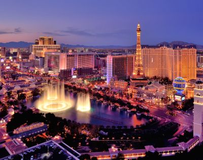 Aerial view of the Las Vegas Strip at dusk. Neon lights illuminate hotels and casinos, including a lit replica Eiffel Tower. A fountain show adds elegance.
