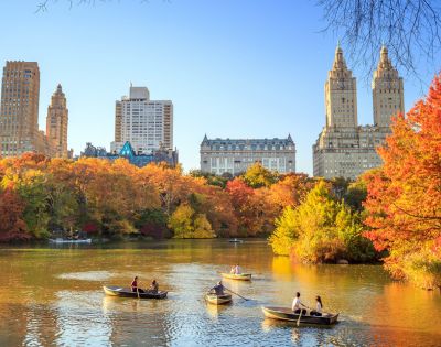Boats with people on a calm lake surrounded by vibrant autumn trees in Central Park, New York City. Tall buildings rise in the background under a clear blue sky.
