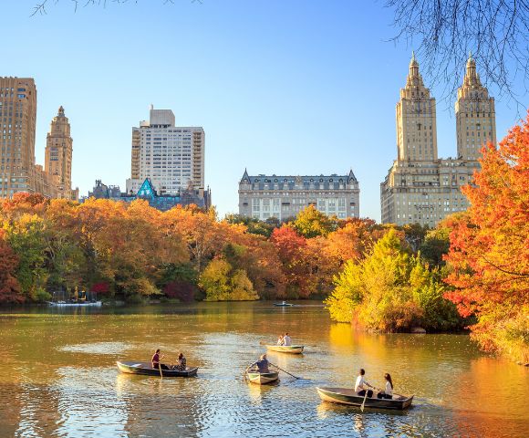 Boats with people on a calm lake surrounded by vibrant autumn trees in Central Park, New York City. Tall buildings rise in the background under a clear blue sky.