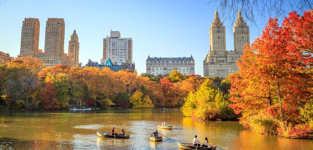 Boats with people on a calm lake surrounded by vibrant autumn trees in Central Park, New York City. Tall buildings rise in the background under a clear blue sky.