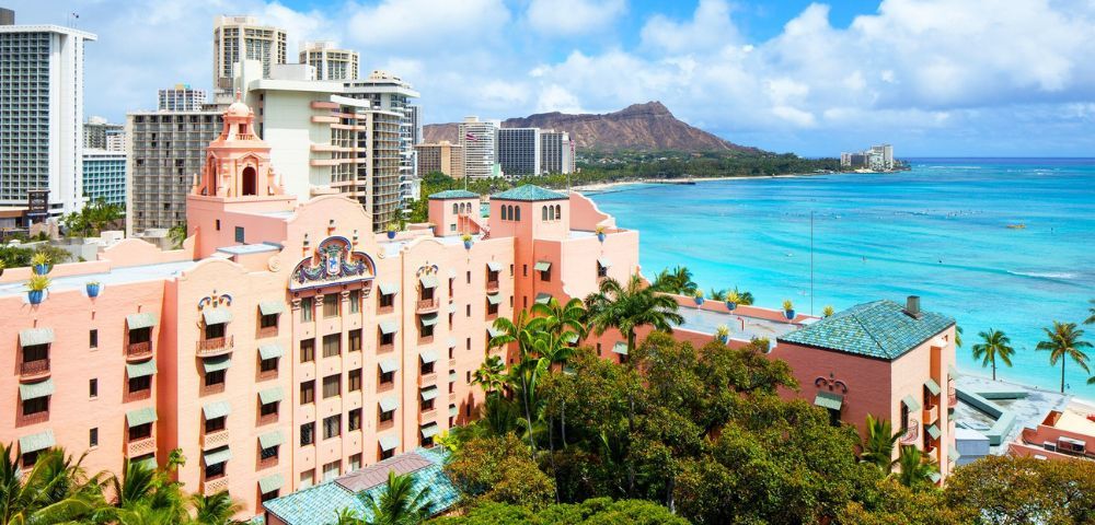 A panoramic view of a pink historic hotel beside palm trees, overlooking turquoise waters and Diamond Head in Honolulu under a blue sky.