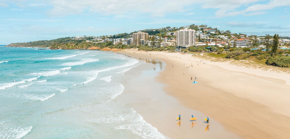 A sunny beach scene with gentle waves, people walking along the shore, and high-rise buildings in the background.