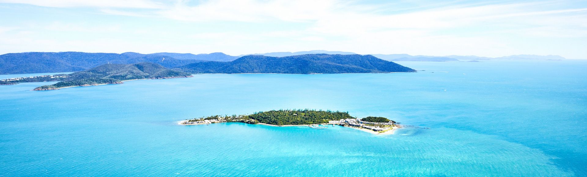 Aerial view of a lush, green island surrounded by vibrant turquoise waters and distant mountains under a bright blue sky.
