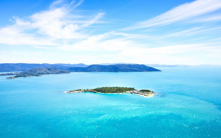 Aerial view of a lush, green island surrounded by vibrant turquoise waters and distant mountains under a bright blue sky.