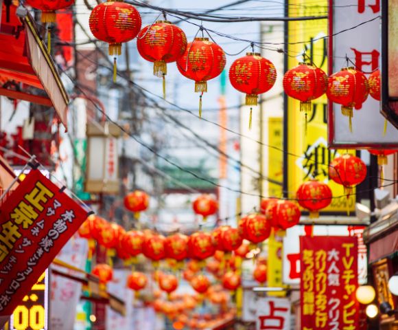A lively street scene decorated with rows of red lanterns and colorful signage, suggesting a festive Asian cultural celebration.