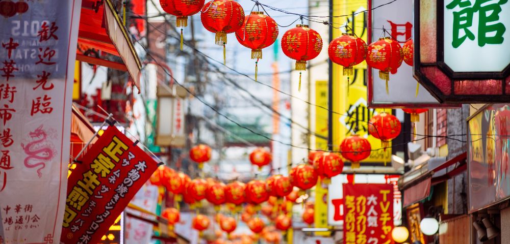 A lively street scene decorated with rows of red lanterns and colorful signage, suggesting a festive Asian cultural celebration.