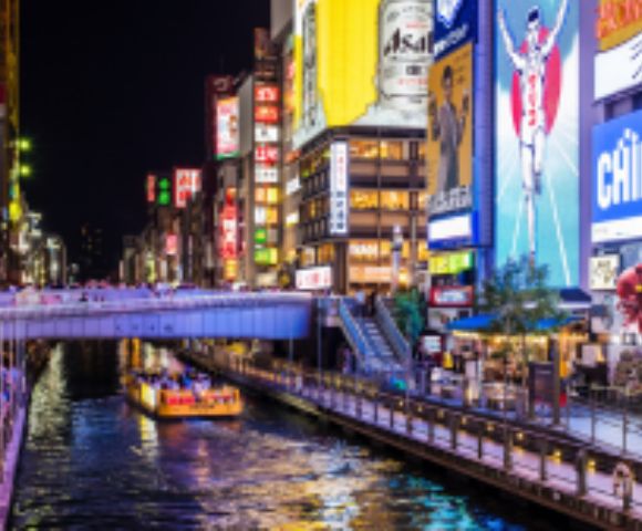 A brightly lit cityscape at night featuring a canal lined with colorful billboards, advertisements, and a boat cruising through the water.
