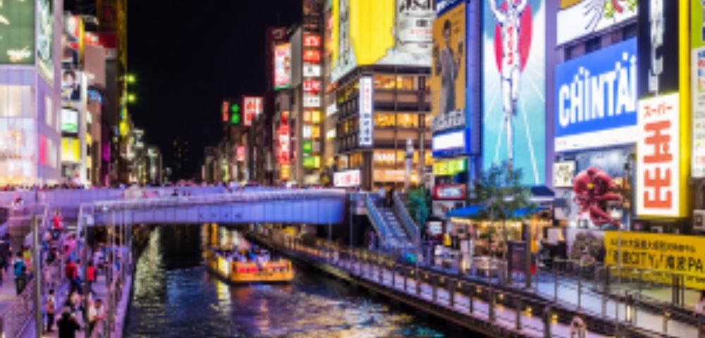 A brightly lit cityscape at night featuring a canal lined with colorful billboards, advertisements, and a boat cruising through the water.