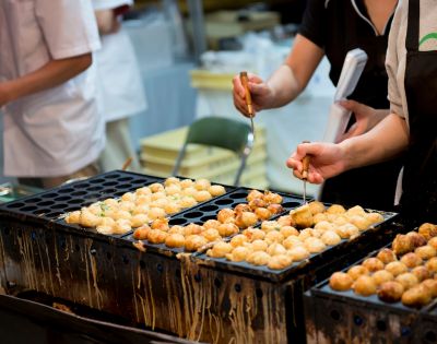 People preparing takoyaki on a specialized grill at a Japanese street food stall.