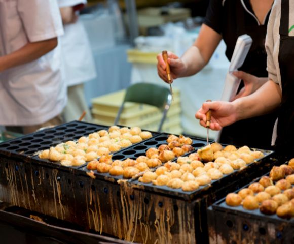 People preparing takoyaki on a specialized grill at a Japanese street food stall.
