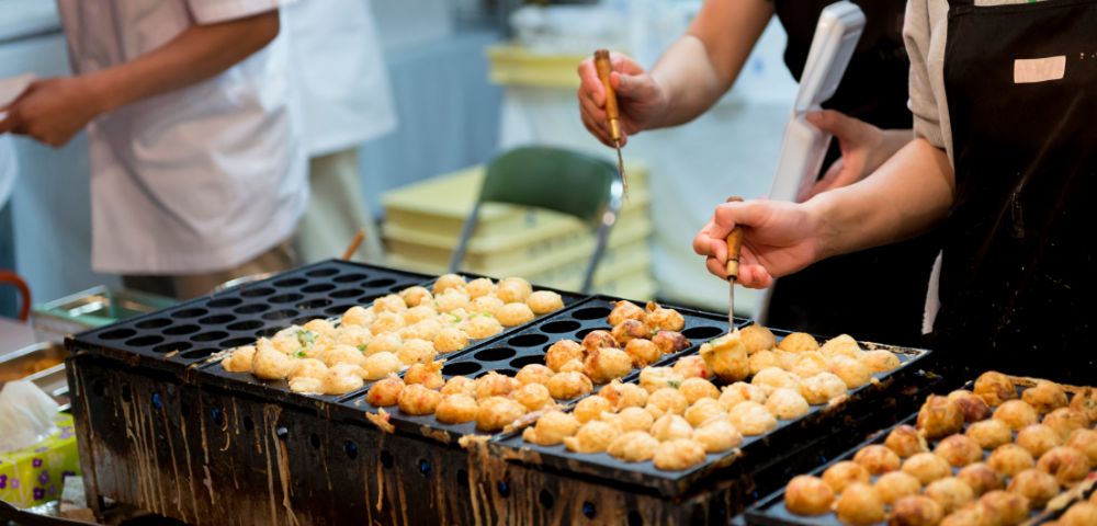 People preparing takoyaki on a specialized grill at a Japanese street food stall.