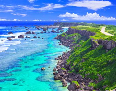 A scenic coastal view featuring turquoise waters, rocky sea formations, and lush green cliffs beneath a bright blue sky.