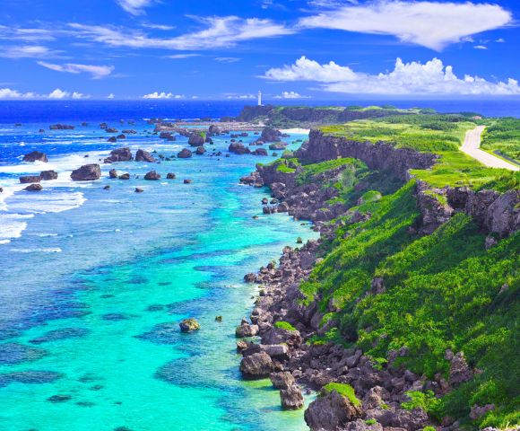 A scenic coastal view featuring turquoise waters, rocky sea formations, and lush green cliffs beneath a bright blue sky.