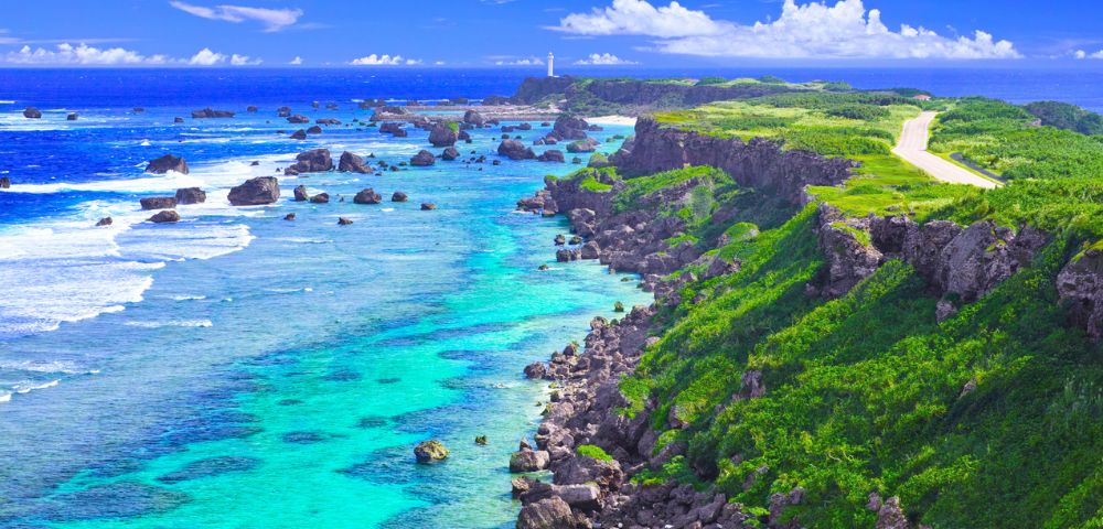 A scenic coastal view featuring turquoise waters, rocky sea formations, and lush green cliffs beneath a bright blue sky.