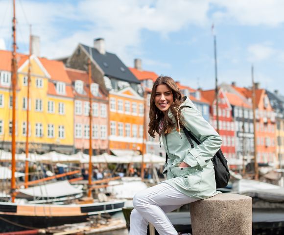 Person sitting on a stone ledge by colorful waterfront buildings and docked boats.