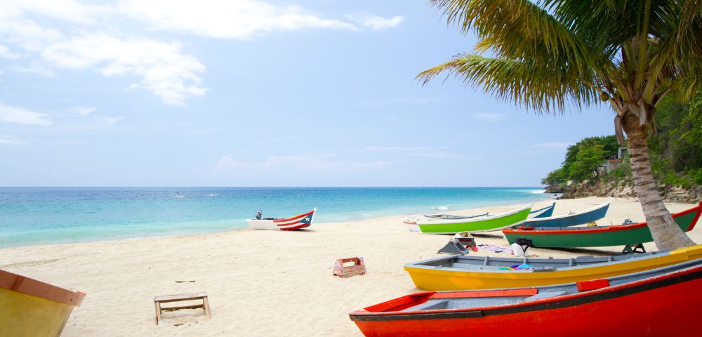 Tropical beach scene with colorful boats on white sand under a palm tree. The turquoise sea and blue sky create a serene, inviting atmosphere.