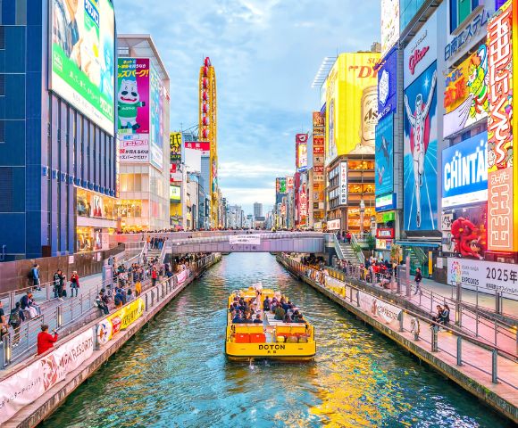 Vibrant urban canal scene with a yellow passenger boat and colorful billboards in Dotonbori, Osaka.