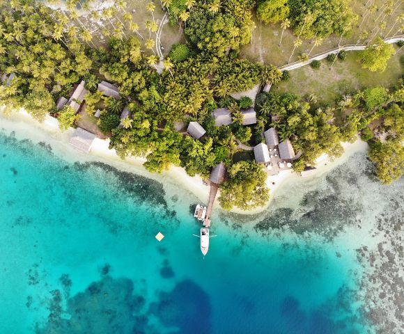 Aerial view of a tropical resort with thatched-roof bungalows near turquoise waters and lush greenery, featuring a docked boat.