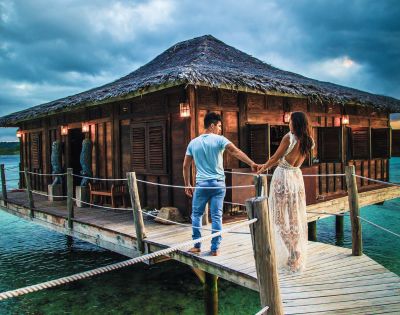 A couple holds hands on a pier leading to a rustic wooden hut, surrounded by calm waters and dramatic clouds above.