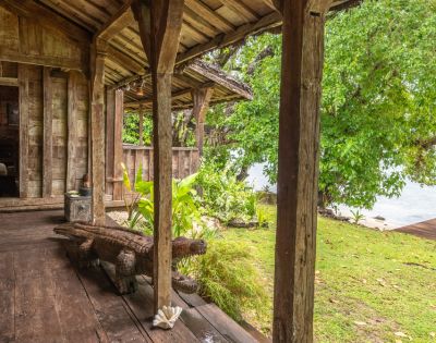 A rustic wooden porch leads to a lush garden and water view, featuring carved animal decor and vibrant greenery.