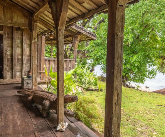 A rustic wooden porch leads to a lush garden and water view, featuring carved animal decor and vibrant greenery.