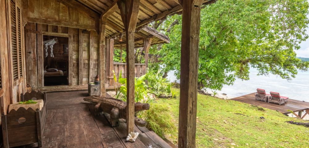 A rustic wooden porch leads to a lush garden and water view, featuring carved animal decor and vibrant greenery.