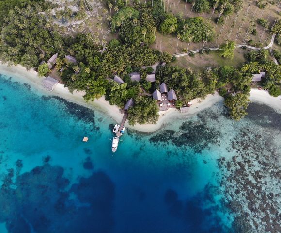 The image features a beautiful tropical beach with a small boat floating on the water near the shore. The boat is located in the middle of the scene, surrounded by a serene environment. In the background, there are several houses and a small village situated on the beach, providing a picturesque view of the coastal area. The houses are scattered along the shoreline, with some closer to the water and others further back on the beach. The overall scene is a perfect representation of a peaceful and idyllic tropical setting.