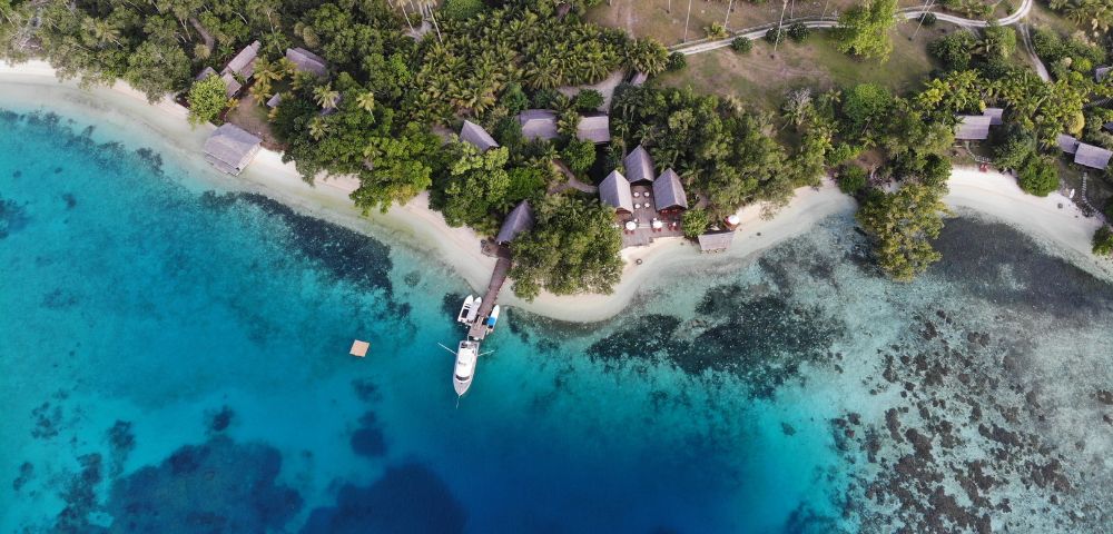 The image features a beautiful tropical beach with a small boat floating on the water near the shore. The boat is located in the middle of the scene, surrounded by a serene environment. In the background, there are several houses and a small village situated on the beach, providing a picturesque view of the coastal area. The houses are scattered along the shoreline, with some closer to the water and others further back on the beach. The overall scene is a perfect representation of a peaceful and idyllic tropical setting.