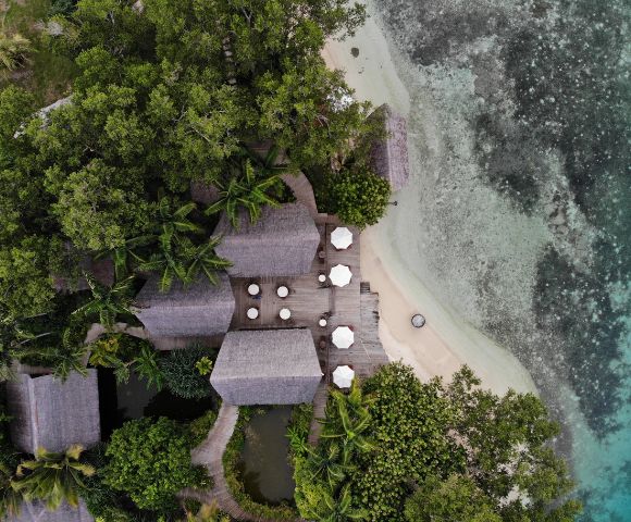 The image features a beautiful tropical beach setting with a wooden deck overlooking the ocean. The deck is surrounded by palm trees, creating a serene and relaxing atmosphere. There are several chairs and dining tables scattered around the deck, providing a comfortable space for people to sit, dine, and enjoy the view. In addition to the chairs and tables, there are multiple umbrellas placed throughout the area, offering shade and protection from the sun. The umbrellas are of various sizes and are positioned at different locations on the deck. The combination of the wooden deck, palm trees