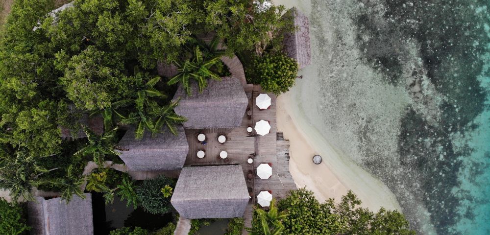 The image features a beautiful tropical beach setting with a wooden deck overlooking the ocean. The deck is surrounded by palm trees, creating a serene and relaxing atmosphere. There are several chairs and dining tables scattered around the deck, providing a comfortable space for people to sit, dine, and enjoy the view. In addition to the chairs and tables, there are multiple umbrellas placed throughout the area, offering shade and protection from the sun. The umbrellas are of various sizes and are positioned at different locations on the deck. The combination of the wooden deck, palm trees