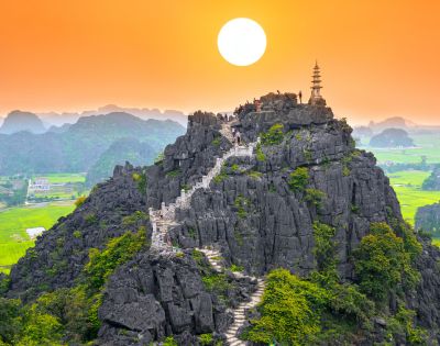A serene sunset over a rocky mountain, featuring a winding staircase leading to a pagoda atop, surrounded by lush green fields.