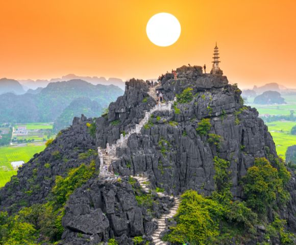 A serene sunset over a rocky mountain, featuring a winding staircase leading to a pagoda atop, surrounded by lush green fields.