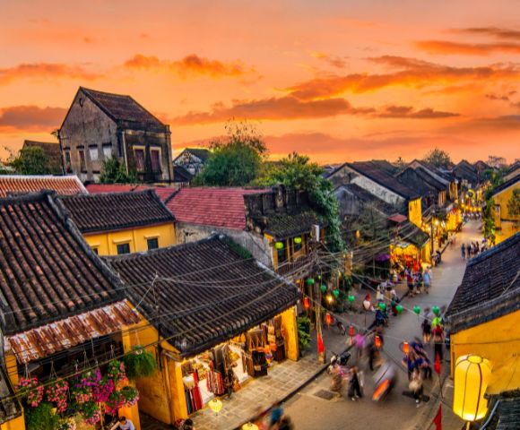 Historic street in Hoi An at sunset, with colorful lanterns, traditional architecture, and lively crowds under a vibrant sky.