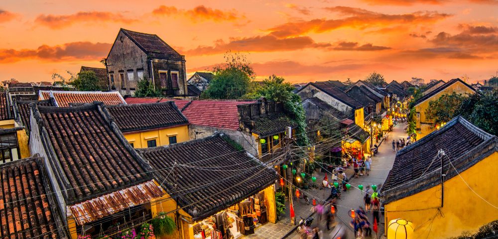Historic street in Hoi An at sunset, with colorful lanterns, traditional architecture, and lively crowds under a vibrant sky.