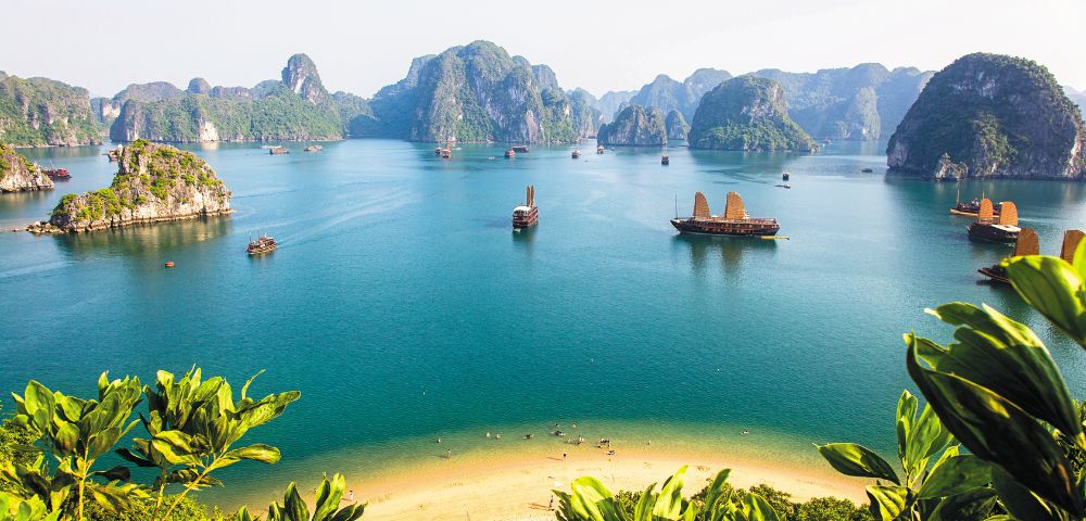 A serene view of Ha Long Bay, with traditional boats sailing through turquoise waters and limestone islands framed by lush greenery.