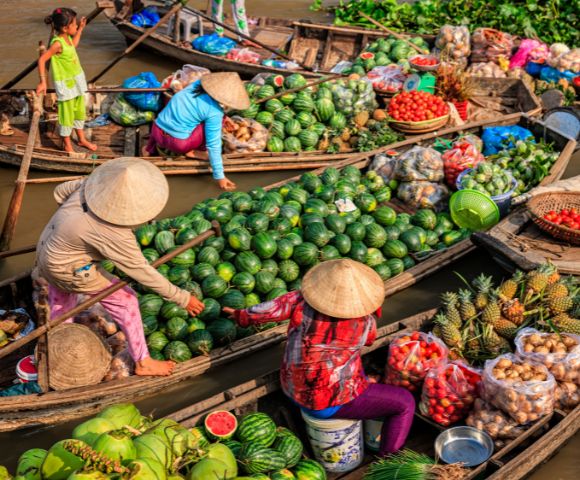 Colorful boats at a bustling market, filled with fresh produce like watermelons, pineapples, and tomatoes, with vendors in traditional hats.