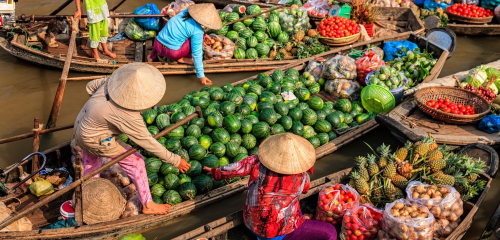 Colorful boats at a bustling market, filled with fresh produce like watermelons, pineapples, and tomatoes, with vendors in traditional hats.