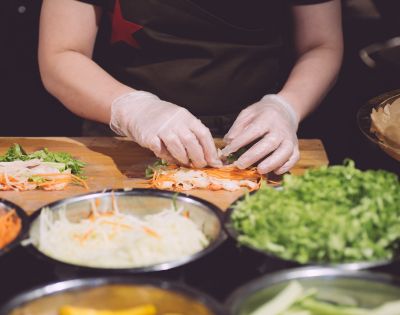 A chef in gloves prepares fresh spring rolls, carefully layering ingredients like shrimp, lettuce, and carrots on a wooden counter.